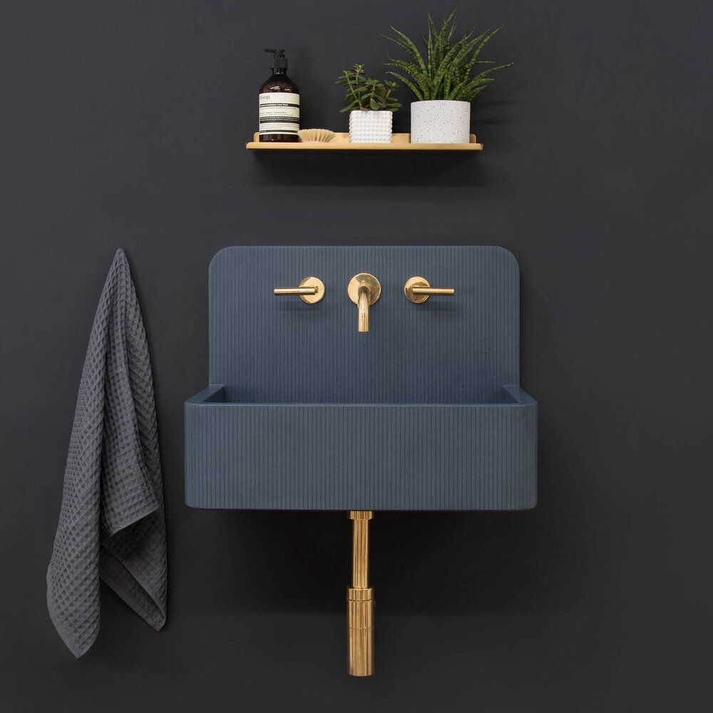 Kast Concrete wall hung elm sink in dark grey with a brushed gold wall mount faucet on a dark grey painted wall with a shelf showing snake plants and organic handsoap