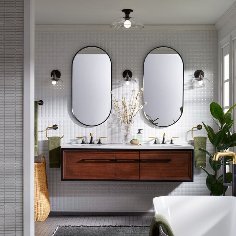 Kohler walnut lodern vanity with double sinks and two capsule mirrors and mixed metal gold and black faucets in a a midcentury modern bathroom green towels freestanding bathroom and three kohler tone sconces