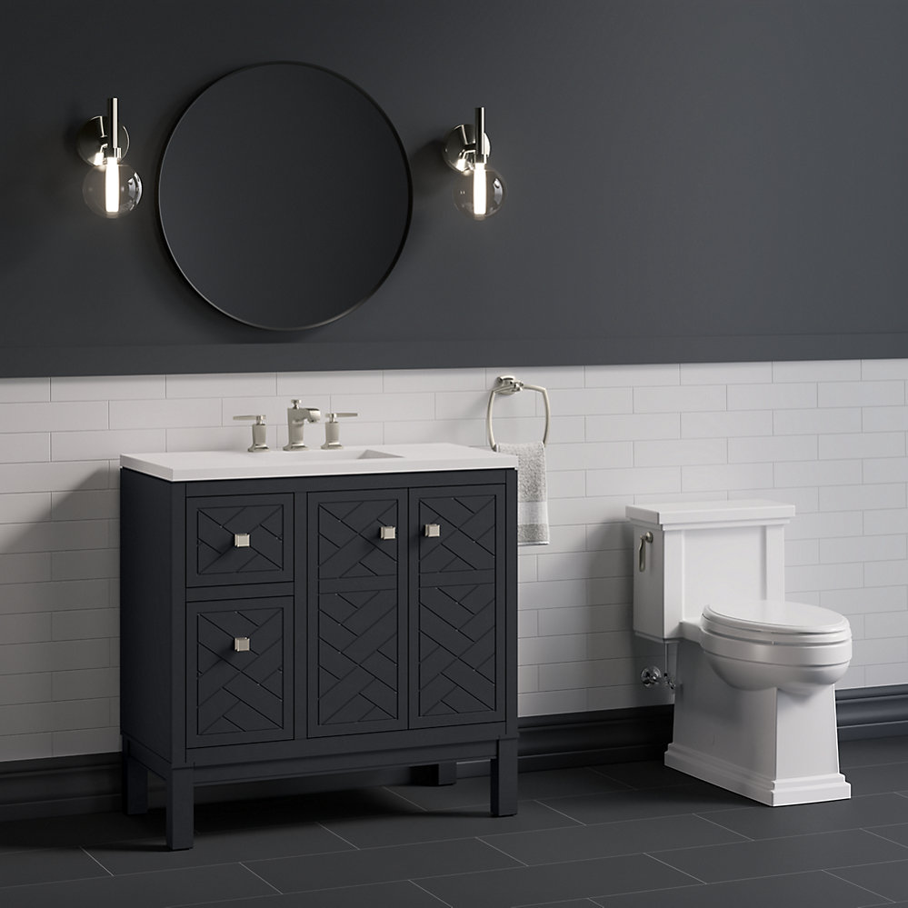 Dark grey kohler bauxline vanity with a round mirror and subway tiles next to a traditional one piece toilet