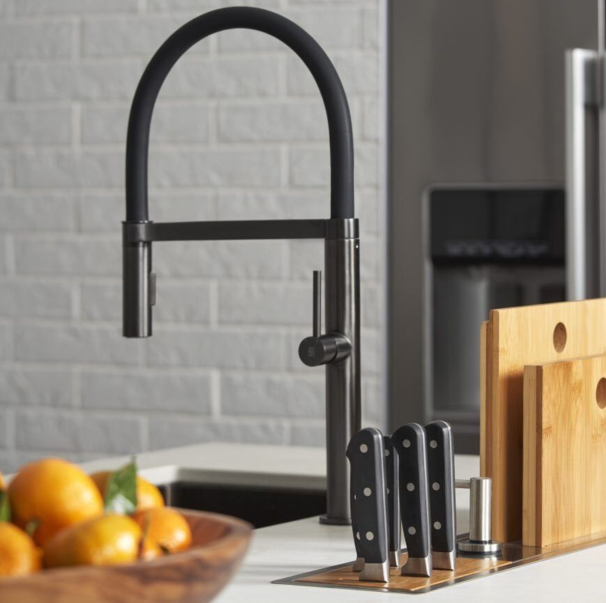 Modern black stainless semi-pro kitchen faucet with workstation trough sink in a loft style kitchen with water dispenser fridge in the background and bowl of fresh oranges in the foreground